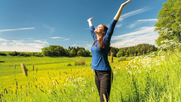 Eine Frau steht auf einem grünen Feld und breitet die Arme aus. Der Himmel ist klar und blau, und die Umgebung ist mit bunten Blumen geschmückt.