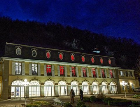 Ein historisches Gebäude mit roten Fenstern, beleuchtet in der Nacht. Im Vordergrund stehen Menschen zwischen gepflegten Gärten und Bänken.