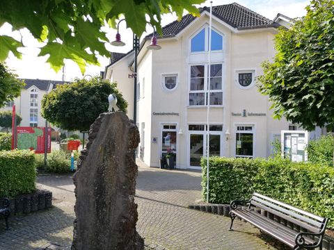 Ein modernes Gebäude mit großen Fenstern und einer Flagge ist im Hintergrund zu sehen. Im Vordergrund sind Bäume und eine Bank platziert.