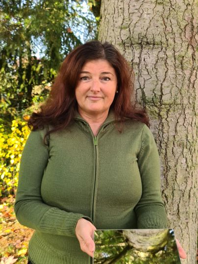 Eine Frau steht vor einem Baum und hält ein Bild mit einer Baumkrone in der Hand. Der Hintergrund ist mit grünem Laub und Bäumen gefüllt.