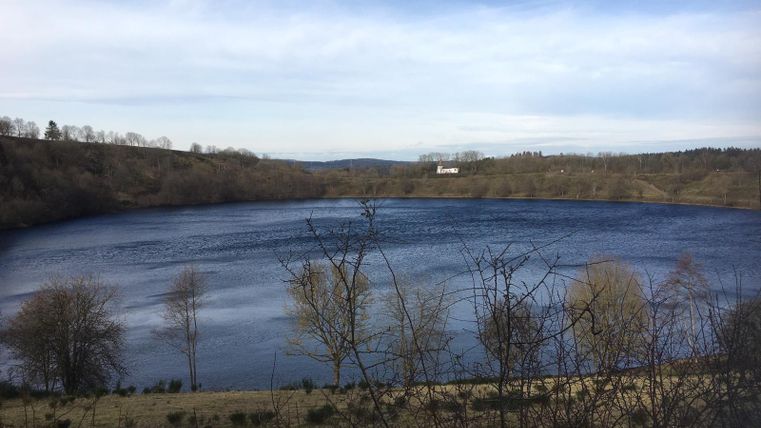 Ein ruhiger See umgeben von sanften Hügeln und kahlen Bäumen. Der Himmel ist klar und der Wasseroberfläche spiegelt das Licht wider.