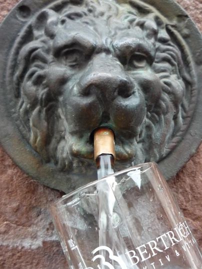 Ein Wasserspender in Form eines Löwenkopfes, aus dem frisches Wasser fließt. Darunter steht ein Glas mit der Aufschrift "Bad Bertrich".