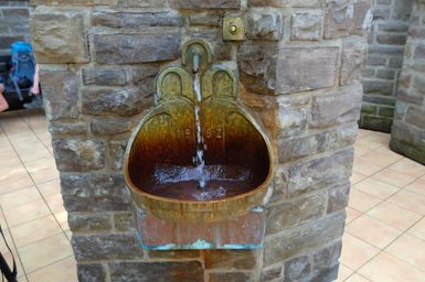 Ein antiker Brunnen aus Metall mit fließendem Wasser. Der Brunnen ist in eine Steinmauer integriert.