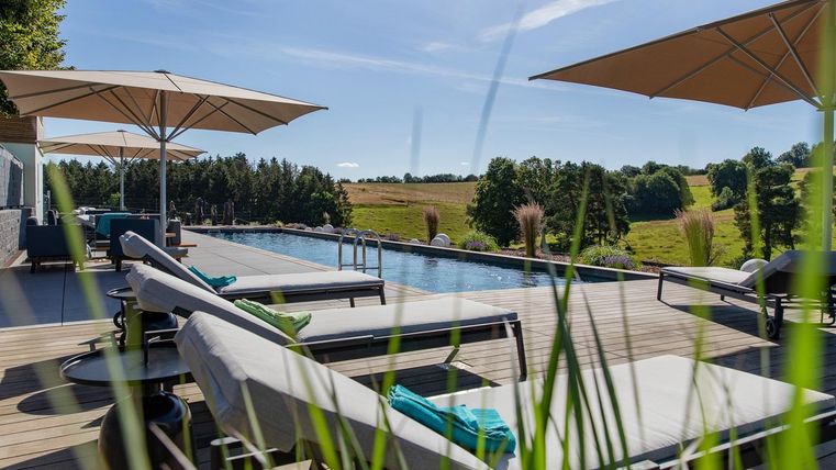 Ein schöner Poolbereich mit Liegen und Sonnenschirmen. Im Hintergrund erstreckt sich eine grüne Landschaft unter blauem Himmel.