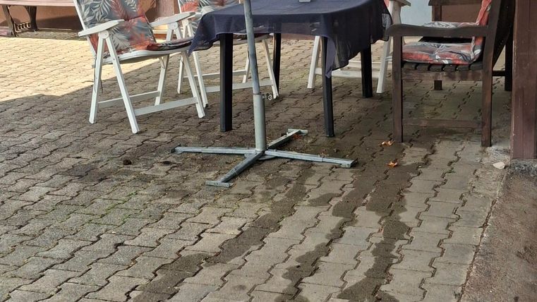 Ein gemütlicher Außenbereich mit einem Tisch und Stühlen unter einem großen Sonnenschirm. Der Boden ist aus Steinplatten und es ist eine ruhige Atmosphäre.