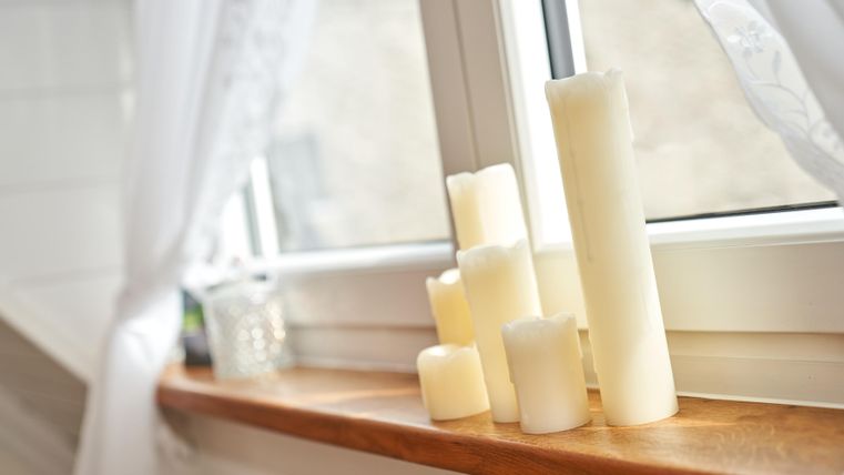 Ein Fenster mit weißen Gardinen und einer Fensterbank, auf der mehrere cremefarbene Kerzen stehen. Die Kerzen sorgen für eine gemütliche Atmosphäre.