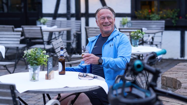 Ein lächelnder Mann sitzt an einem Tisch im Freien und genießt ein Bier. Der Tisch ist mit einer kleinen Pflanze dekoriert und im Hintergrund sind leere Stühle zu sehen.
