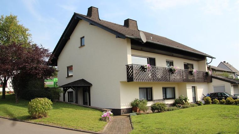 Ein großes, zweistöckiges Haus mit einem Balkon und Blumen. Der Garten ist gepflegt und es gibt einige Bäume und Sträucher.