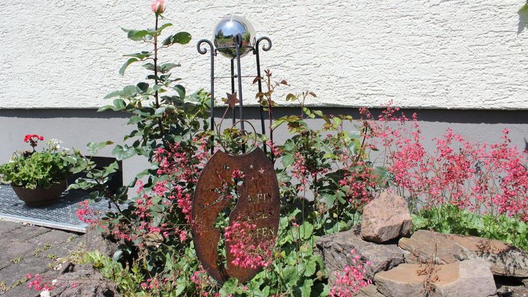 Eine dekorative Gartenstelle mit einer Metalltafel und vielen bunten Blumen. Im Hintergrund ist eine helle Wand zu sehen.