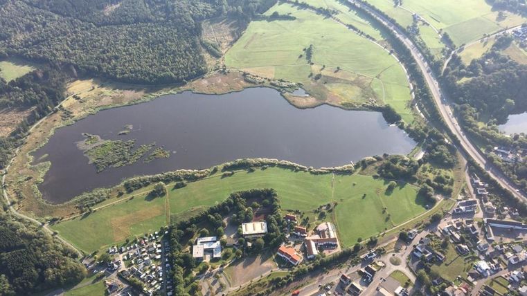 Ein weitläufiges Landschaftsbild mit einem ruhigen See, umgeben von grünen Wiesen und Bäumen. Im Vordergrund sind einige Gebäude und Straßen sichtbar.