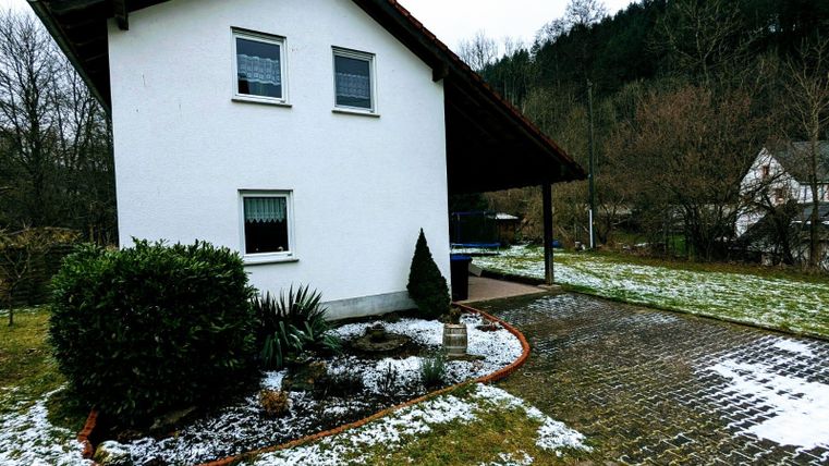 Ein weißes Haus mit einem roten Dach und einem kleinen Vorgarten. Der Boden ist teilweise mit Schnee bedeckt und es gibt Bäume im Hintergrund.