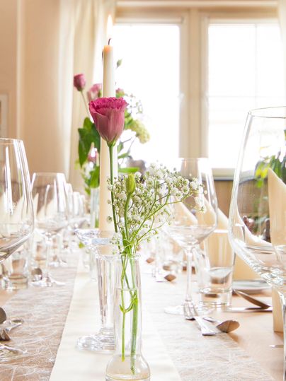Ein festlich gedeckter Tisch mit Gläsern und Blumen in einer Vase. Im Hintergrund ist ein Kerzenlicht und ein heller Raum zu sehen.