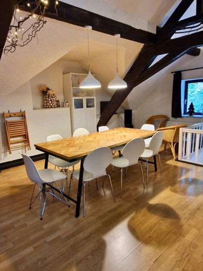 Ein gemütlicher Essbereich mit einem langen Holztisch und weißen Stühlen. Der Raum hat hohe Decken und sichtbare Balken, sowie warmes Licht von Hängeleuchten.