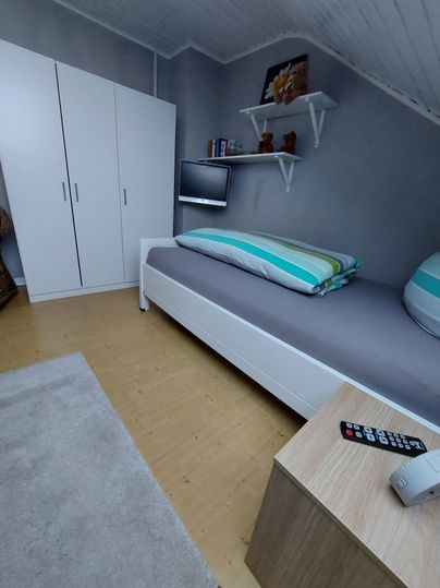 Ein einfaches Zimmer mit einem Einzelbett, einem Fernseher und einem Kleiderschrank. Der Boden ist aus Holz und ein kleiner Tisch steht neben dem Bett.