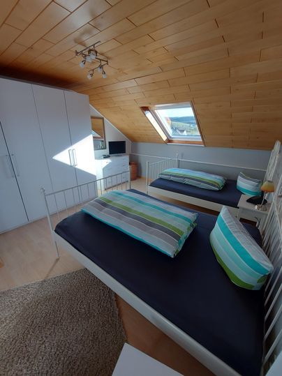 Ein gemütliches Schlafzimmer mit zwei Einzelbetten und bunten Kissen. Große Fenster sorgen für viel Tageslicht und einen Blick nach draußen.