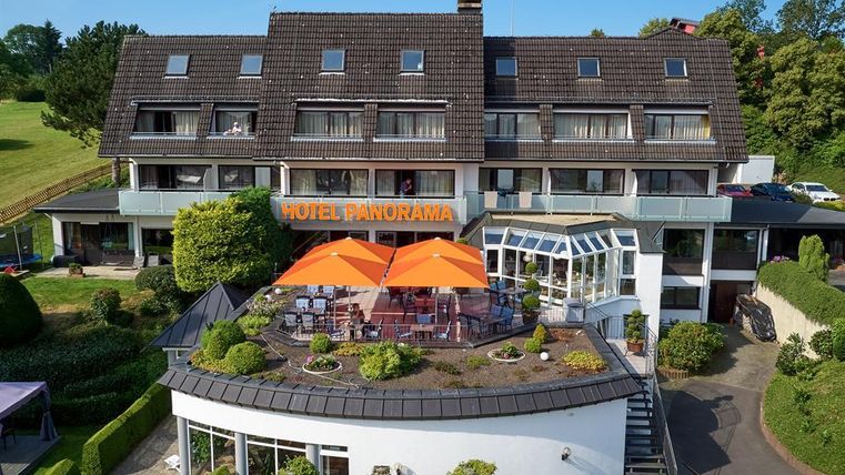 Ein modernes Hotel mit dem Namen „Hotel Panorama“ und einer einladenden Terrasse. Die Terrasse ist mit orangefarbenen Sonnenschirmen und grünen Pflanzen dekoriert.