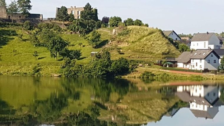 Eine malerische Landschaft mit einem ruhigen See und grünen Hügeln. Im Hintergrund sind historische Gebäude und kleinere Häuser zu sehen.