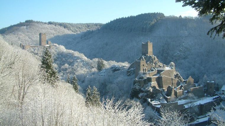 Eine winterliche Landschaft mit schneebedeckten Hügeln und einer alten Burgruine. Der Himmel ist klar und die Bäume sind von Frost bedeckt.