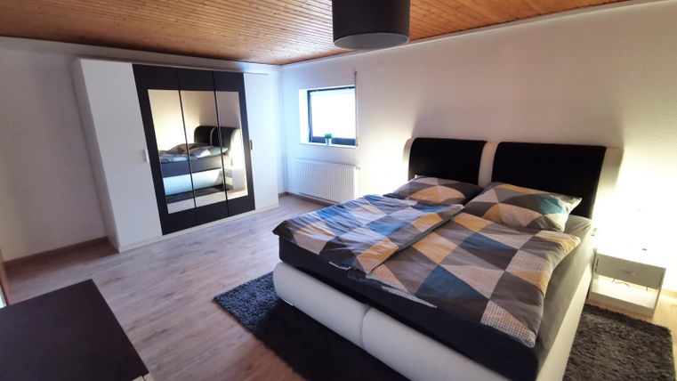 Ein modernes Schlafzimmer mit einem großen Bett und einem Spiegelschrank. Der Raum hat Holzboden und sanfte Beleuchtung.
