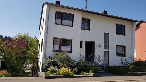 Ein zweigeschossiges, helles Wohnhaus mit weißen Wänden. Der Vorgarten ist grün und blühend, mit verschiedenen Pflanzen.