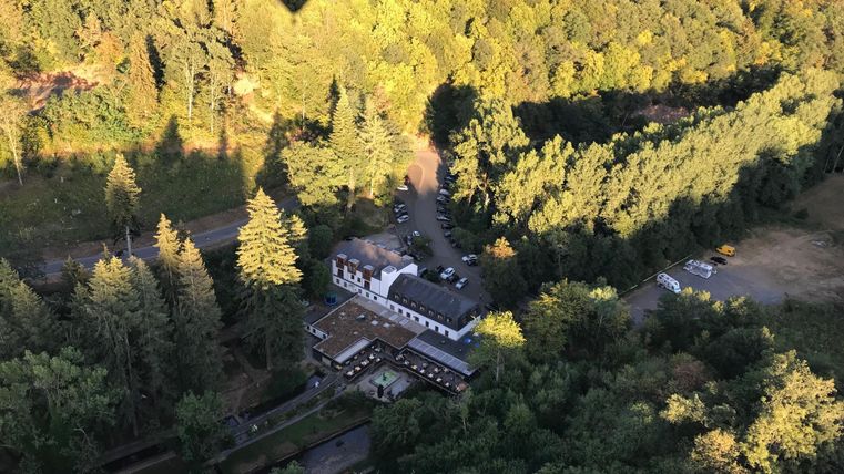 Eine Luftaufnahme eines ruhigen Waldgebiets mit einem Gebäude im Vordergrund. Im Hintergrund zeichnen sich Bäume und der Schatten eines Heißluftballons ab.