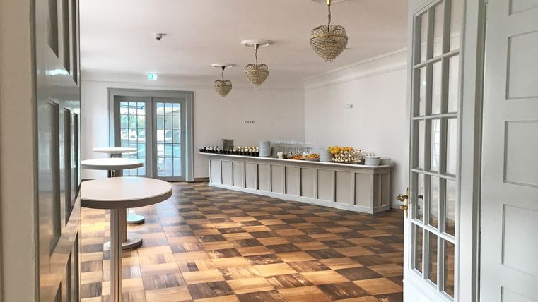 Ein helles, modernes Zimmer mit einem Holzfußboden. Auf einer langen Anrichte sind verschiedene Speisen und Getränke bereitgestellt.