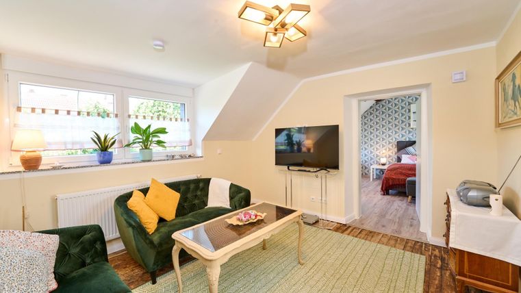 Ein gemütliches Wohnzimmer mit einem grünen Sofa und einem Tisch. Der Raum hat große Fenster und ist hell erleuchtet.
