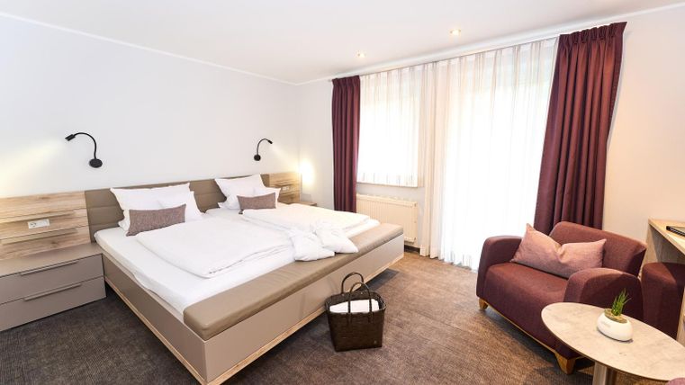 Ein modernes Hotelzimmer mit einem großen Doppelbett und einem gemütlichen Sessel. Es gibt viele Fenster, die für Tageslicht sorgen, und eine stilvolle Dekoration.