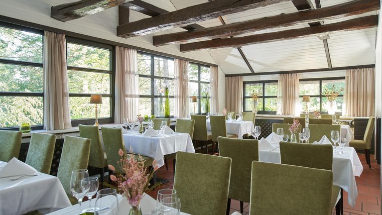 Ein eleganter Restaurantbereich mit grünem Mobiliar und Tischen, die mit weißen Tischdecken gedeckt sind. Große Fenster bieten Blick auf die grüne Umgebung.