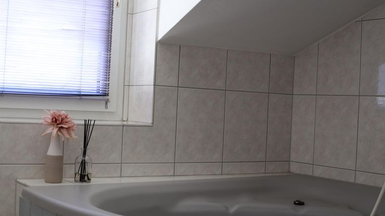 Ein modernes Badezimmer mit einer Eckbadewanne und hellen Fliesen. Ein Fenster mit einem blauen Rollos und dekorativen Elementen vervollständigt den Raum.