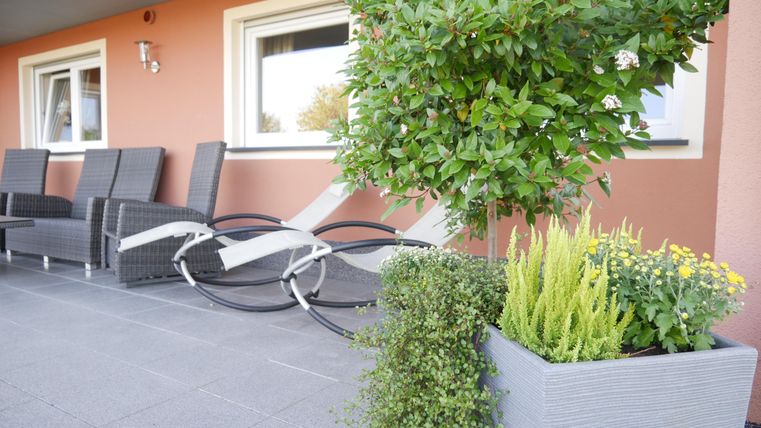 Ein gemütlicher Außenbereich mit Blick auf eine Terrasse. Es gibt bequeme Stühle, Pflanzen und einen schön gestalteten Blumenkasten.