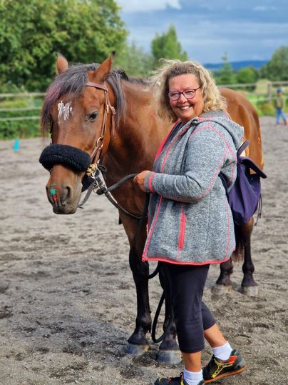 Eine Frau steht neben einem Pferd auf einem reitbaren Platz. Es ist eine freundliche Atmosphäre mit grünem Hintergrund.