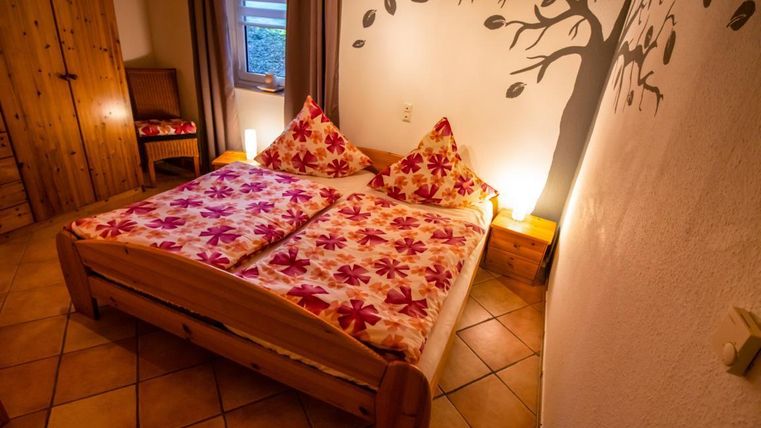Ein gemütliches Schlafzimmer mit einem Doppelbett und bunten Blumenmustern auf der Bettwäsche. An der Wand ist ein Baum-Motiv und es gibt warme Lichtquellen, die eine einladende Atmosphäre schaffen.