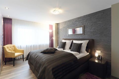 Ein modernes Schlafzimmer mit einem großen Bett und stilvollen Möbeln. Ein bequemer Sessel und viel Tageslicht schaffen eine einladende Atmosphäre.