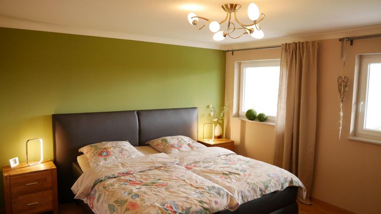 Ein gemütliches Schlafzimmer mit einem Doppelbett und bunten Bettbezügen. An den Wänden sind grüne und beige Farbtöne, und die Fenster lassen viel Tageslicht herein.