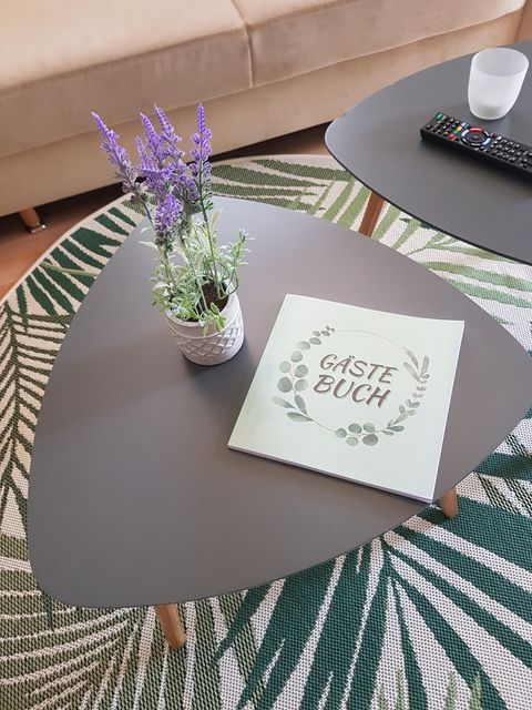 Ein stilvoller Couchtisch mit einem Gästebuch und einer blühenden Pflanze. Im Hintergrund sieht man ein gemütliches Sofa und einen dekorativen Teppich.
