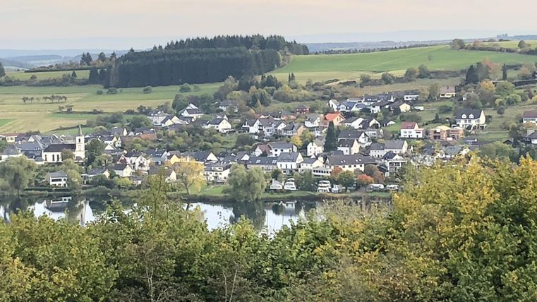 Eine malerische Landschaft mit einem kleinen Dorf am Fluss. Umgeben von grünen Hügeln und Bäumen.