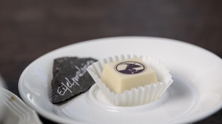Eine exquisite Praline auf einem weißen Teller, umgeben von einer eleganten Papierschnur. Daneben liegt ein schwarzes Stück Tafel mit einer handschriftlichen Notiz.
