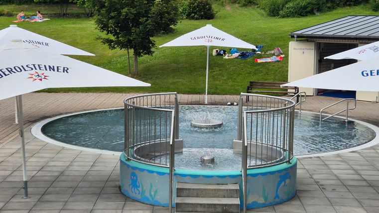 Ein kleiner Whirlpool mit Wasser und einem Eingang aus Stufen. Rundherum sind Sonnenschirme und eine grüne Wiese im Hintergrund.