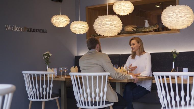 Ein modernes Restaurant mit zwei Gästen, die am Tisch sitzen und sich unterhalten. Sie sind von stilvollen Lampen und einer eleganten Einrichtung umgeben.
