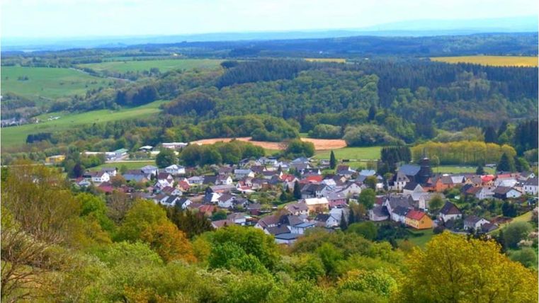 Eine malerische Aussicht auf ein kleines Dorf umgeben von grünen Wäldern und Feldern. Der Himmel ist klar und die Landschaft ist idyllisch.