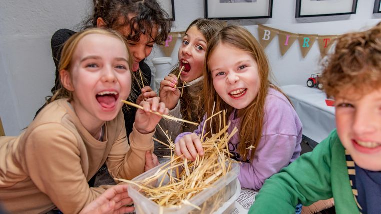 Eine Gruppe von Kindern lächelt und spielt zusammen. Sie halten Stäbchen und stehen vor einer Box mit Strohhalmen.