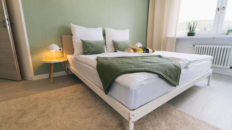 Ein modernes Schlafzimmer mit einem weißen Bett und grünen Kissen. Der Raum hat eine ruhige Atmosphäre und viel Tageslicht.