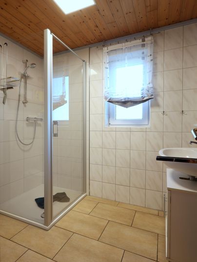 Ein modernes Badezimmer mit einer Duschkabine und einem Waschbecken. Die Wände sind gefliest, und es gibt ein Fenster mit Vorhängen.