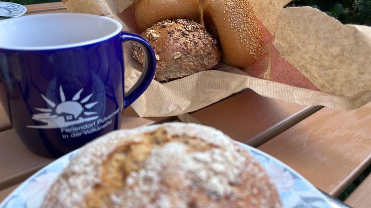 Eine Tasse Kaffee und frische Brötchen auf einem Tisch. Im Hintergrund sind weitere Brötchen in einer Papierverpackung sichtbar.