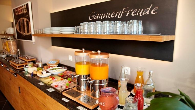 Ein einladendes Frühstücksbuffet mit verschiedenen Speisen und Getränken. Im Vordergrund stehen zwei Saftspender und frische Zutaten auf einem Tisch.