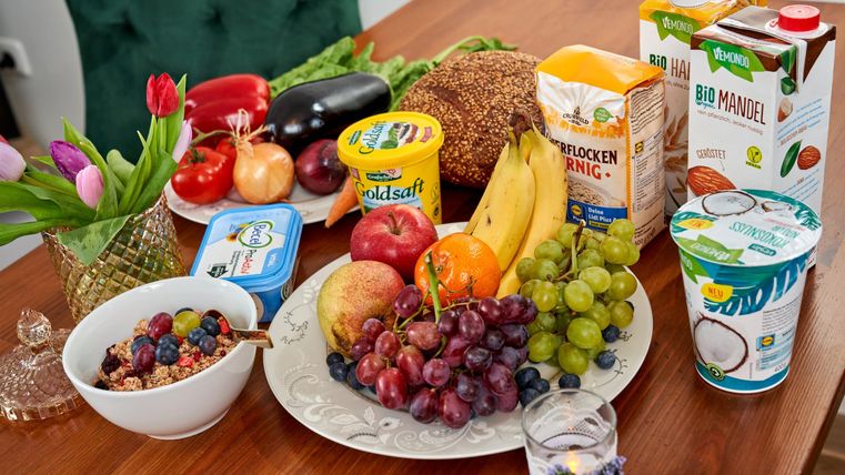 Ein Tisch mit einer bunten Auswahl an frischem Obst, darunter Trauben, Äpfel und Bananen. Neben dem Obst stehen verschiedene Getränke und Snacks bereit.