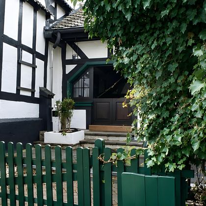 Ein hübsches Fachwerkhaus mit grünen Fensterrahmen und einer geschützten Eingangstür. Der ebene Garten ist mit einer grünen Holzlattenzaun umgeben.