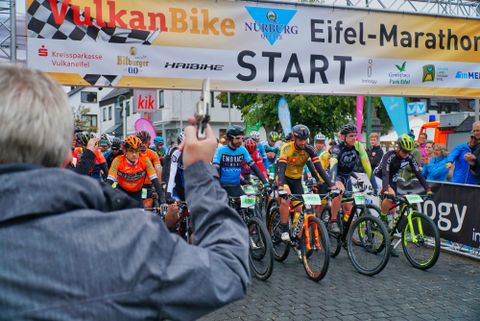 Eine Gruppe von Radfahrern steht am Start des Eifel-Marathons. Im Hintergrund sieht man ein Startbanner und Zuschauer, die auf das Rennen warten.