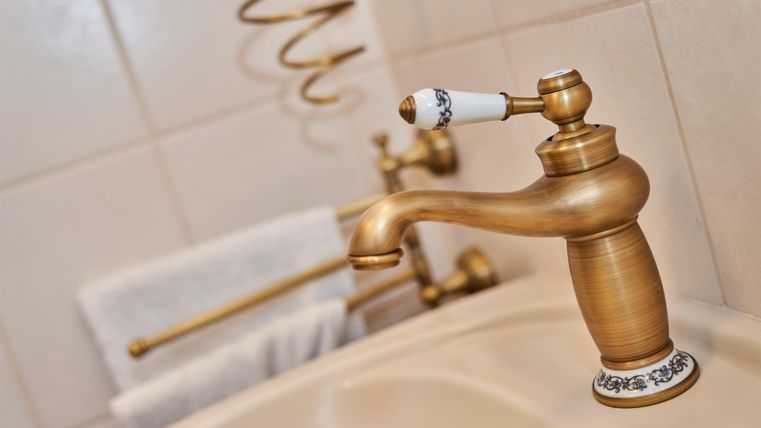 Ein eleganter Wasserhahn aus goldenem Metall in einem Badezimmer. Im Hintergrund sind Handtücher und eine dekorative Stange zu sehen.